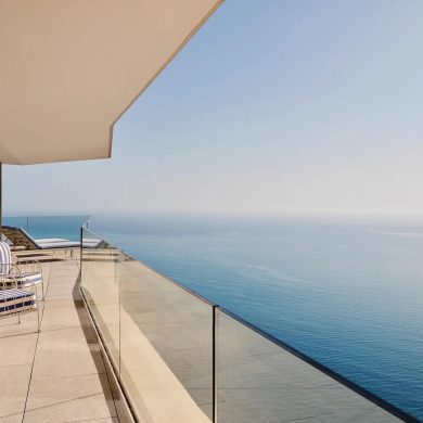 Modern balcony overlooking the ocean with glass railing and blue‑striped lounge chairs along a tiled terrace.