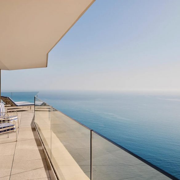 Modern balcony overlooking the ocean with glass railing and blue‑striped lounge chairs along a tiled terrace.
