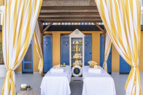 Outdoor spa cabanas with yellow-and-white striped drapes, blue doors, and white massage tables with towels and toiletries on a sunny deck