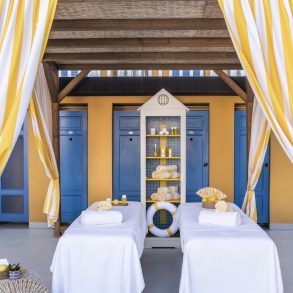 Outdoor spa cabanas with yellow-and-white striped drapes, blue doors, and white massage tables with towels and toiletries on a sunny deck