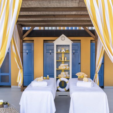 Outdoor spa cabanas with yellow-and-white striped drapes, blue doors, and white massage tables with towels and toiletries on a sunny deck