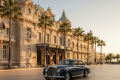 Classic dark-blue vintage car parked on a tiled promenade in front of a grand ornate beige hotel with palm trees on either side.