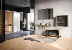 Modern bathroom showroom with grey textured wall, gold faucet, wall mirror, and floating countertop against light wood flooring.