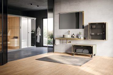 Modern bathroom showroom with grey textured wall, gold faucet, wall mirror, and floating countertop against light wood flooring.