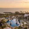 Sunset over a resort pool area with palm trees and ocean in the distance.