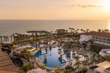 Sunset over a resort pool area with palm trees and ocean in the distance.