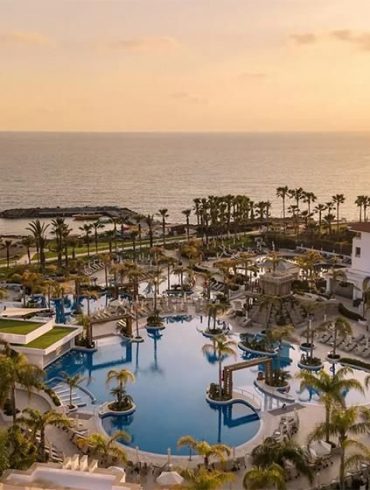 Sunset over a resort pool area with palm trees and ocean in the distance.