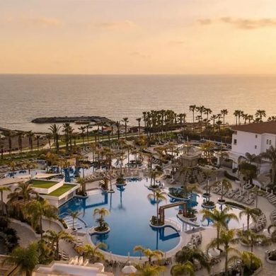 Sunset over a resort pool area with palm trees and ocean in the distance.