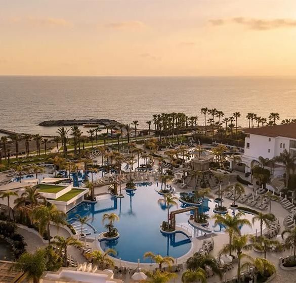 Sunset over a resort pool area with palm trees and ocean in the distance.
