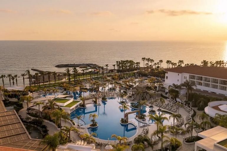 Sunset over a resort pool area with palm trees and ocean in the distance.