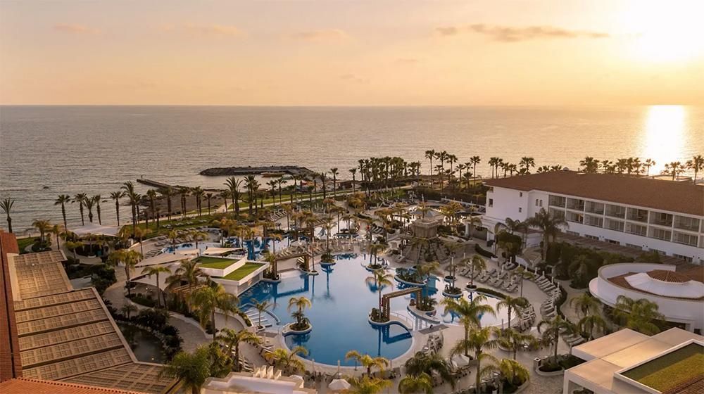Sunset over a resort pool area with palm trees and ocean in the distance.