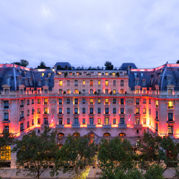 Elegant Parisian-style building with ornate balconies, illuminated in pink and orange lights at dusk, with trees in the foreground