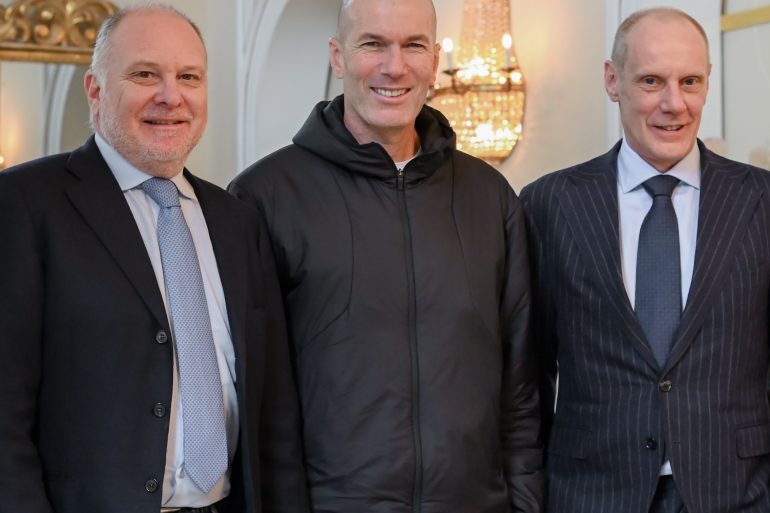 Three men in business attire smiling for a group photo indoors, left in a dark suit, center in a black jacket, right in a pinstripe suit.