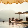 Poolside scene with lounge chairs, beige and blue umbrellas, and people swimming by the edge with mountains in the distance.