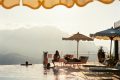 Poolside scene with lounge chairs, beige and blue umbrellas, and people swimming by the edge with mountains in the distance.