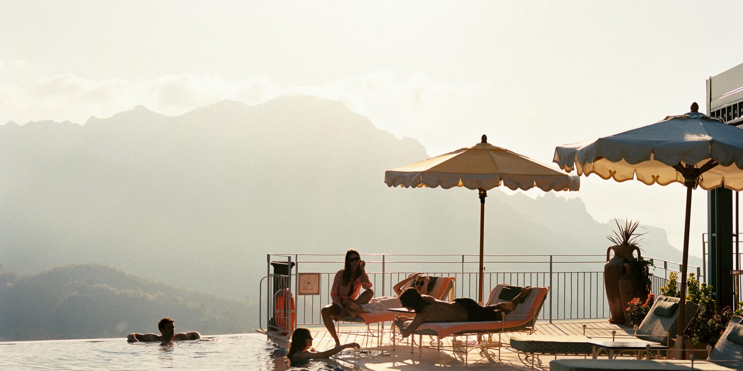 Poolside scene with lounge chairs, beige and blue umbrellas, and people swimming by the edge with mountains in the distance.