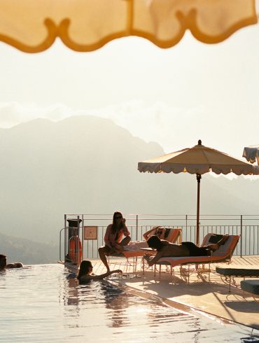 Poolside scene with lounge chairs, beige and blue umbrellas, and people swimming by the edge with mountains in the distance.