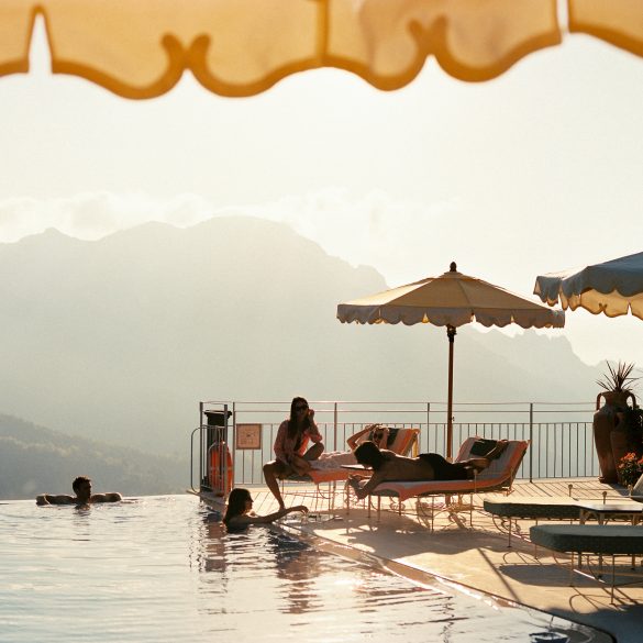 Poolside scene with lounge chairs, beige and blue umbrellas, and people swimming by the edge with mountains in the distance.