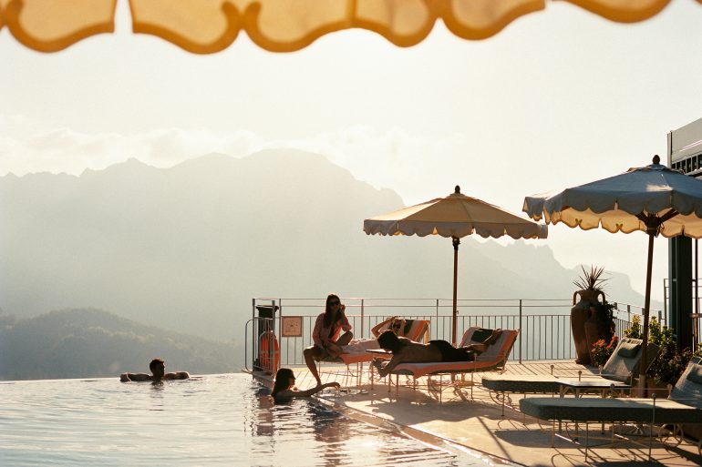 Poolside scene with lounge chairs, beige and blue umbrellas, and people swimming by the edge with mountains in the distance.