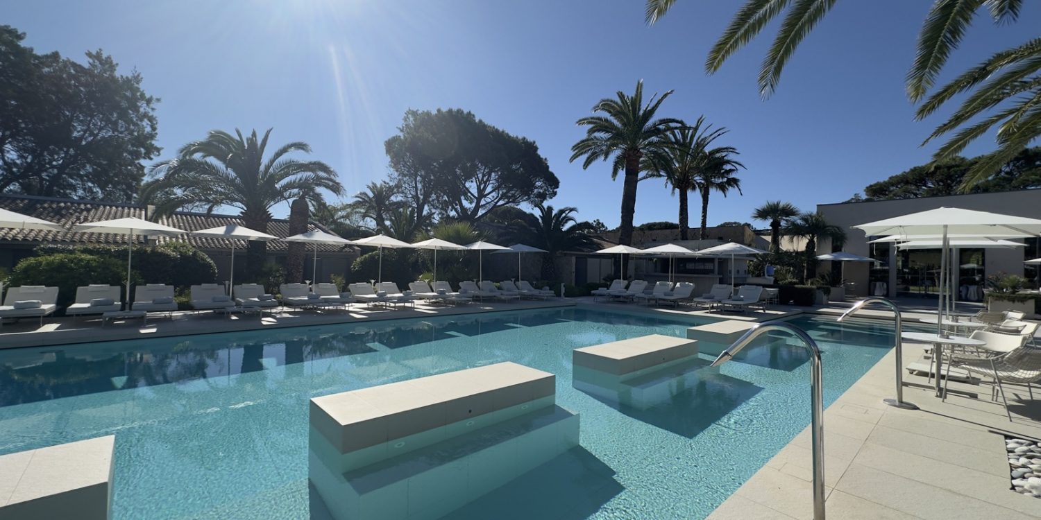 Sunlit resort pool with turquoise water, white loungers and umbrellas along the deck, palm trees in the background.