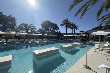 Sunlit resort pool with turquoise water, white loungers and umbrellas along the deck, palm trees in the background.