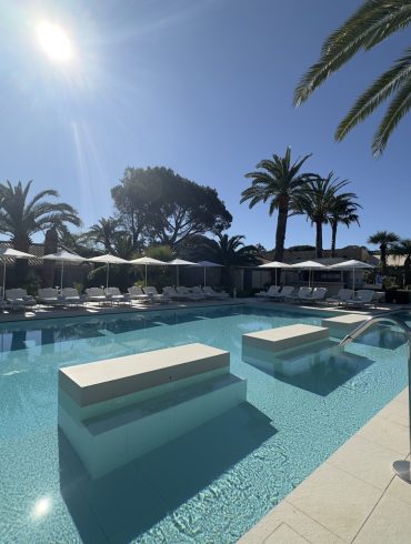 Sunlit resort pool with turquoise water, white loungers and umbrellas along the deck, palm trees in the background.