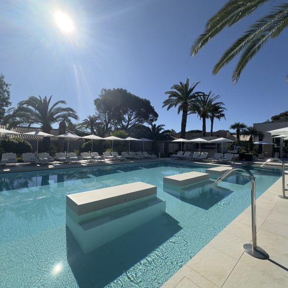 Sunlit resort pool with turquoise water, white loungers and umbrellas along the deck, palm trees in the background.