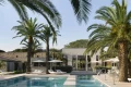 Luxurious outdoor resort pool area with palm trees, white lounge chairs, and umbrellas beside a modern villa.