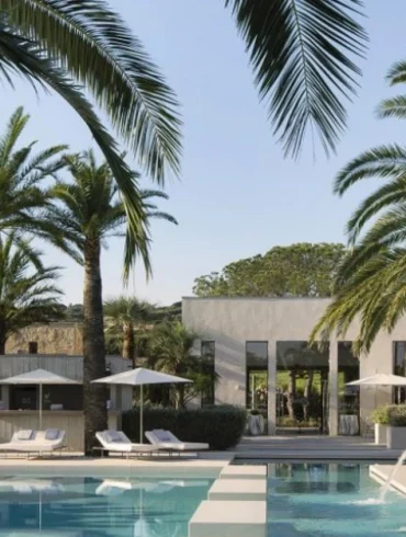 Luxurious outdoor resort pool area with palm trees, white lounge chairs, and umbrellas beside a modern villa.
