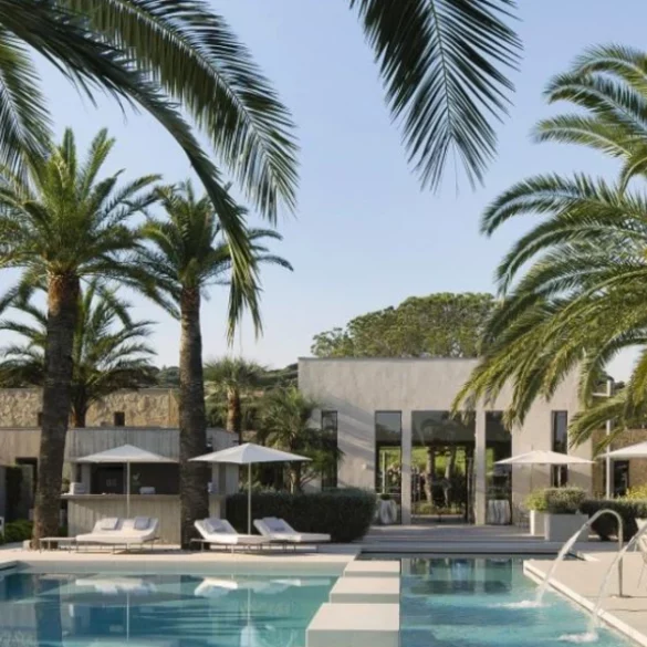 Luxurious outdoor resort pool area with palm trees, white lounge chairs, and umbrellas beside a modern villa.