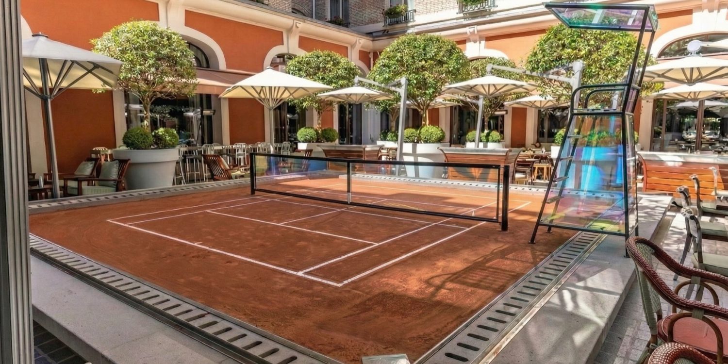 Outdoor courtyard with a small clay tennis court, surrounded by orange walls, white arches, and potted trees.
