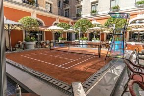 Outdoor courtyard with a small clay tennis court, surrounded by orange walls, white arches, and potted trees.