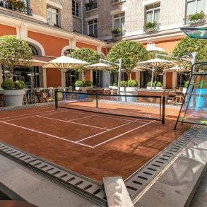 Outdoor courtyard with a small clay tennis court, surrounded by orange walls, white arches, and potted trees.