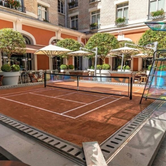 Outdoor courtyard with a small clay tennis court, surrounded by orange walls, white arches, and potted trees.