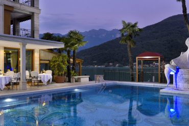 Poolside view of a luxury hotel at dusk with illuminated dining tables on the deck and a sculpted fountain beside palm trees overlooking a lake and mountains in the background.