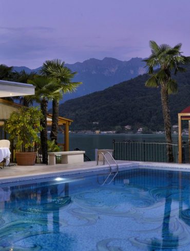 Poolside view of a luxury hotel at dusk with illuminated dining tables on the deck and a sculpted fountain beside palm trees overlooking a lake and mountains in the background.