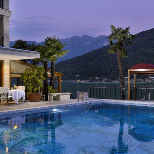 Poolside view of a luxury hotel at dusk with illuminated dining tables on the deck and a sculpted fountain beside palm trees overlooking a lake and mountains in the background.