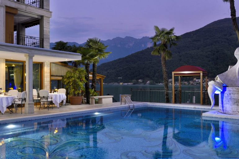 Poolside view of a luxury hotel at dusk with illuminated dining tables on the deck and a sculpted fountain beside palm trees overlooking a lake and mountains in the background.