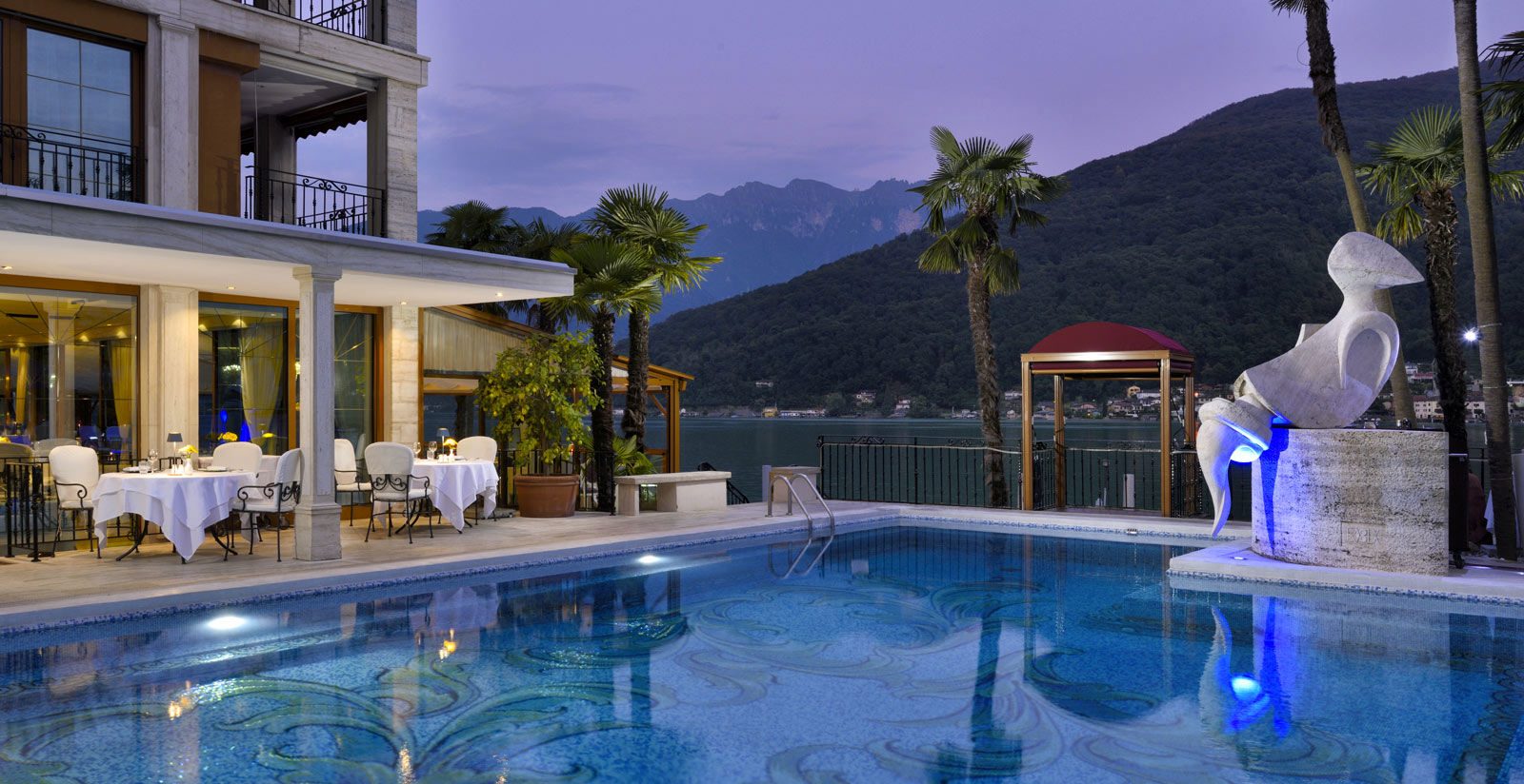 Poolside view of a luxury hotel at dusk with illuminated dining tables on the deck and a sculpted fountain beside palm trees overlooking a lake and mountains in the background.
