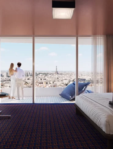 Couple stands on a balcony in a modern hotel room, overlooking a city skyline with the Eiffel Tower in the distance.