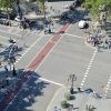 A busy urban intersection viewed from above with a red bike lane running through the center, pedestrians on sidewalks, cyclists ahead, and parked motorcycles along the curb.