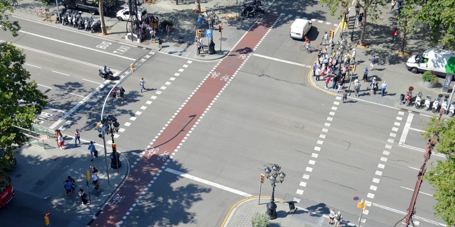 A busy urban intersection viewed from above with a red bike lane running through the center, pedestrians on sidewalks, cyclists ahead, and parked motorcycles along the curb.