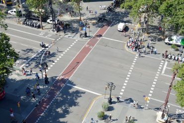 A busy urban intersection viewed from above with a red bike lane running through the center, pedestrians on sidewalks, cyclists ahead, and parked motorcycles along the curb.