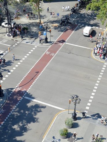 A busy urban intersection viewed from above with a red bike lane running through the center, pedestrians on sidewalks, cyclists ahead, and parked motorcycles along the curb.