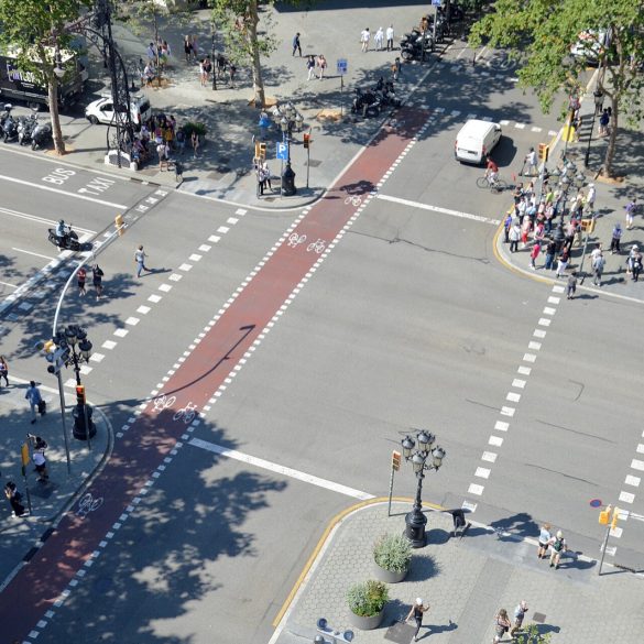 A busy urban intersection viewed from above with a red bike lane running through the center, pedestrians on sidewalks, cyclists ahead, and parked motorcycles along the curb.