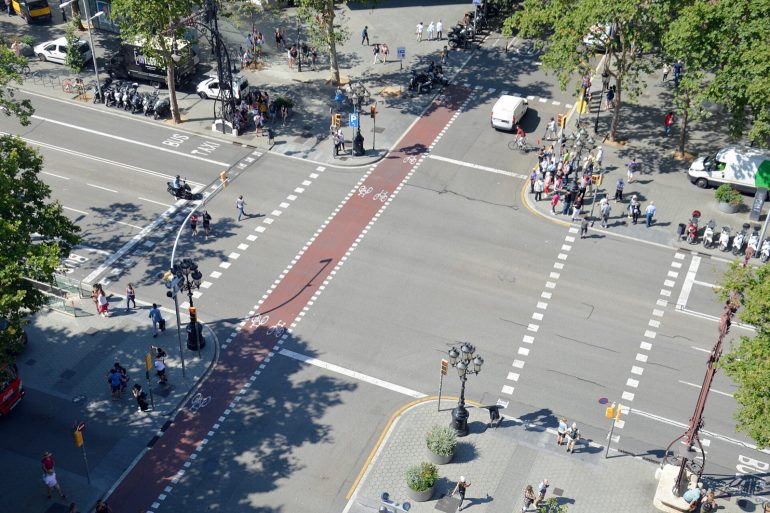 A busy urban intersection viewed from above with a red bike lane running through the center, pedestrians on sidewalks, cyclists ahead, and parked motorcycles along the curb.