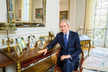 Man in a dark suit sits at a polished wooden desk in a richly decorated room with gold-framed mirrors and portrait photos on the desk.
