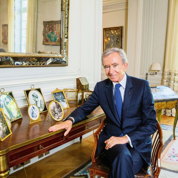 Man in a dark suit sits at a polished wooden desk in a richly decorated room with gold-framed mirrors and portrait photos on the desk.