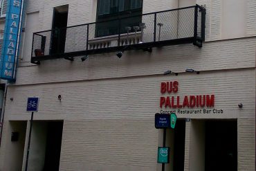 White brick building with a black metal balcony above the entrance; red letters spell 'BUS PALLADIUM' and a 'Concert Restaurant Bar Club' sign on the façade.