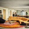 Mid‑century living room with a curved orange velvet sofa, black chair, and round coffee tables on a warm gradient rug by mirrored walls.